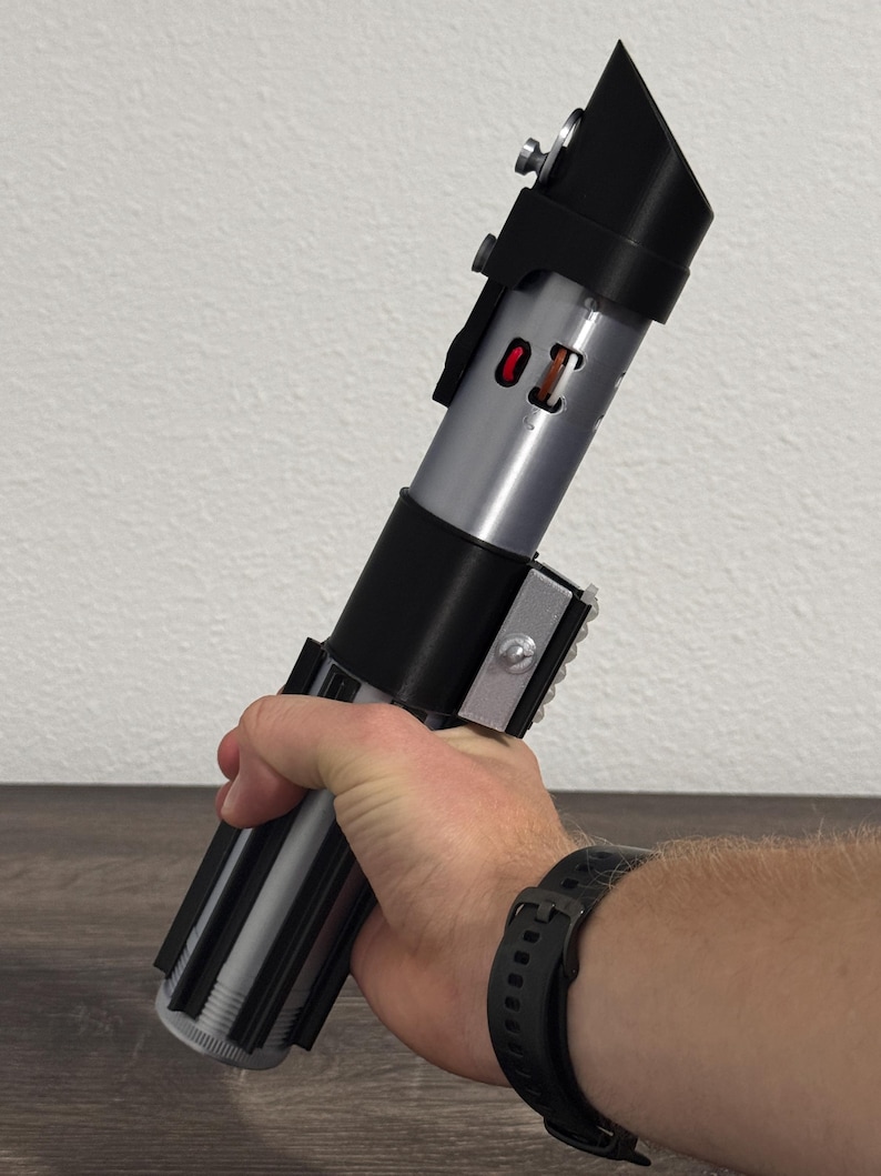 Pu&ograve; includere: Un'elica di spada laser argento e nera con un pulsante rosso e la scritta "01". L'elica ha un'impugnatura nera e un corpo argentato. La spada laser &egrave; tenuta in mano.