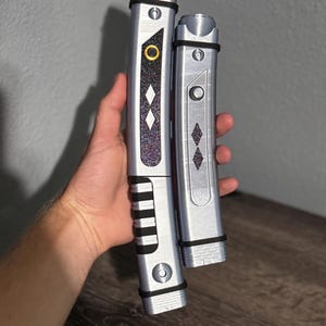 Pu&ograve; includere: Due spade laser argentate, una pi&ugrave; grande e una pi&ugrave; piccola, con dettagli neri e forme a diamante decorative. La spada laser pi&ugrave; grande ha una parte superiore dorata e un'impugnatura nera. La spada laser pi&ugrave; piccola ha una parte superiore argentata. Entrambe hanno un elemento di design circolare.