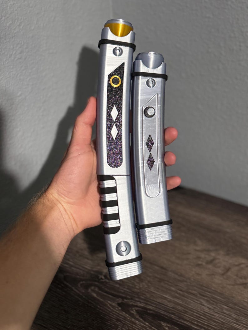 Pu&ograve; includere: Due spade laser argentate, una pi&ugrave; grande e una pi&ugrave; piccola, con dettagli neri e forme a diamante decorative. La spada laser pi&ugrave; grande ha una parte superiore dorata e un'impugnatura nera. La spada laser pi&ugrave; piccola ha una parte superiore argentata. Entrambe hanno un elemento di design circolare.