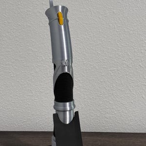 May include: A silver lightsaber prop with a black grip and a yellow button. The lightsaber is standing upright on a black base. The hilt has a textured appearance, and a silver blade-like element extends from the top.