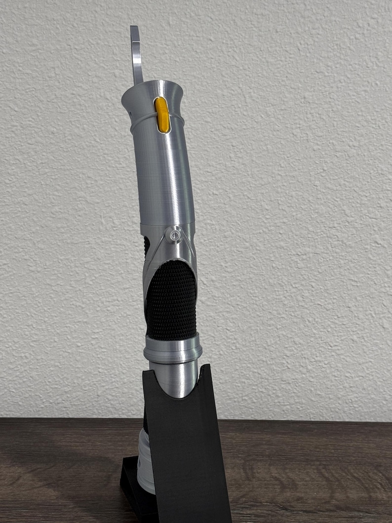 May include: A silver lightsaber prop with a black grip and a yellow button. The lightsaber is standing upright on a black base. The hilt has a textured appearance, and a silver blade-like element extends from the top.