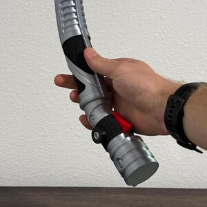 May include: A silver and black lightsaber prop with a red button and textured grip. The prop is held in a person's hand, showcasing its design and details. The lightsaber has a curved design and a cylindrical base.