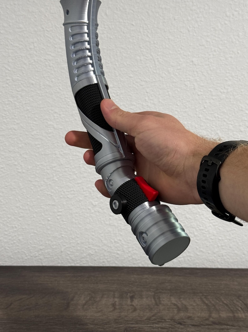 May include: A silver and black lightsaber prop with a red button and textured grip. The prop is held in a person's hand, showcasing its design and details. The lightsaber has a curved design and a cylindrical base.