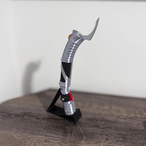 May include: A silver and black lightsaber with a curved blade, resting on a black stand. The hilt features a red button and a yellow accent. The lightsaber is displayed on a wooden surface against a white background.