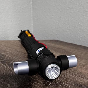 May include: A black lightsaber prop with silver detailing and a bright LED light. The hilt has a complex design with red wiring and a yellow component. The lightsaber is on a wooden surface.