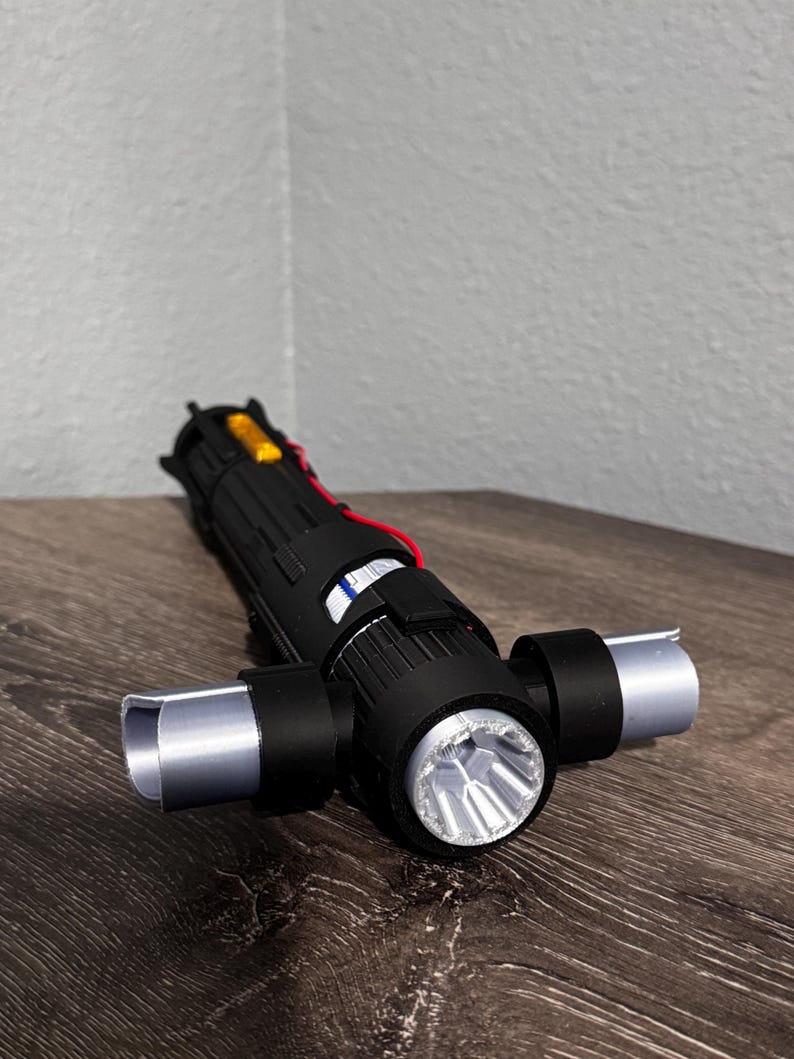 May include: A black lightsaber prop with silver detailing and a bright LED light. The hilt has a complex design with red wiring and a yellow component. The lightsaber is on a wooden surface.