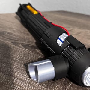May include: A black and silver lightsaber prop with exposed wiring and a silver emitter. The prop has a cylindrical handle with detailed ridges and a silver end cap. A yellow component is visible near the top.