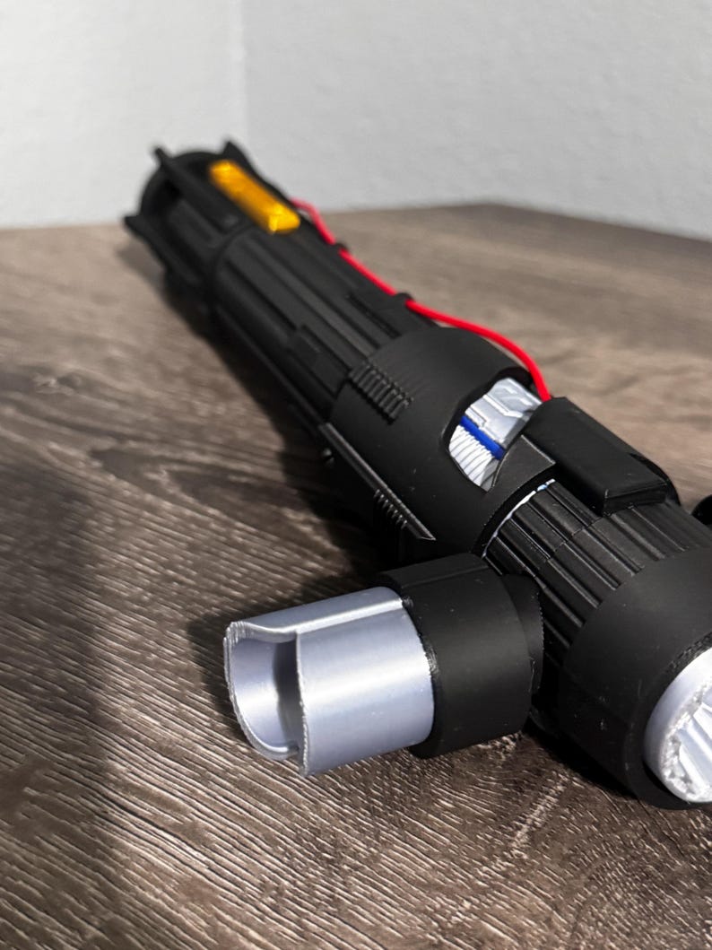 May include: A black and silver lightsaber prop with exposed wiring and a silver emitter. The prop has a cylindrical handle with detailed ridges and a silver end cap. A yellow component is visible near the top.