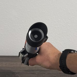 Pu&ograve; includere: Un'elica di spada laser nera e argento tenuta in mano. La spada laser ha un'impugnatura cilindrica con un emettitore nero e un corpo argentato. La mano indossa un orologio nero.