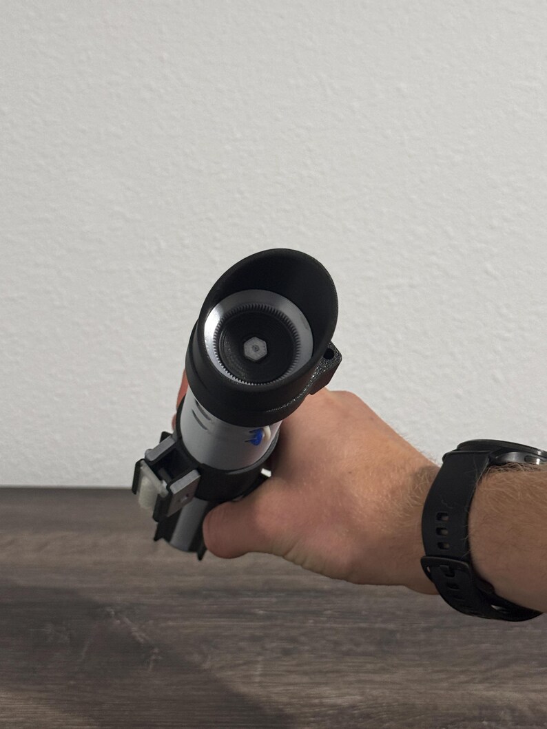 Pu&ograve; includere: Un'elica di spada laser nera e argento tenuta in mano. La spada laser ha un'impugnatura cilindrica con un emettitore nero e un corpo argentato. La mano indossa un orologio nero.