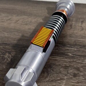 Puede incluir: Un accesorio de sable de luz plateado y negro con una pieza de acento amarilla y marr&oacute;n. El accesorio tiene un dise&ntilde;o cil&iacute;ndrico con un mango detallado y una secci&oacute;n superior con una tapa plateada. El sable de luz es una r&eacute;plica de un arma de ciencia ficci&oacute;n popular.