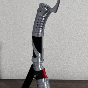 May include: A silver and black lightsaber prop with a curved design, featuring a red button and a black grip. The prop is displayed on a black stand, showcasing its intricate details and futuristic aesthetic.