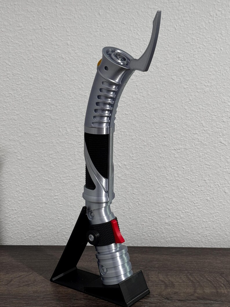 May include: A silver and black lightsaber prop with a curved design, featuring a red button and a black grip. The prop is displayed on a black stand, showcasing its intricate details and futuristic aesthetic.