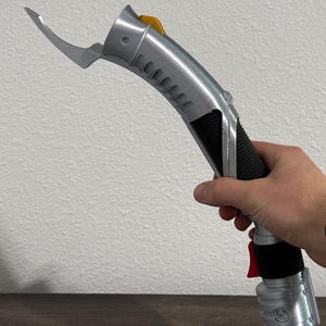 May include: A silver and black lightsaber with a curved blade extension and a yellow button. The handle has a black grip and a red button near the base. The lightsaber is held against a white wall.