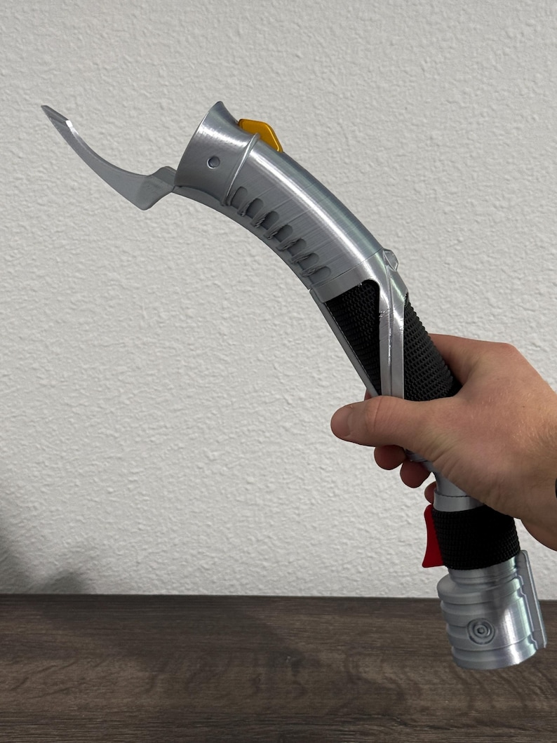 May include: A silver and black lightsaber with a curved blade extension and a yellow button. The handle has a black grip and a red button near the base. The lightsaber is held against a white wall.