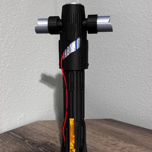 May include: A black lightsaber with silver crossguard blades and a red wire detail. The hilt has a cylindrical design with a gold and red accent. The lightsaber is displayed on a wooden surface.