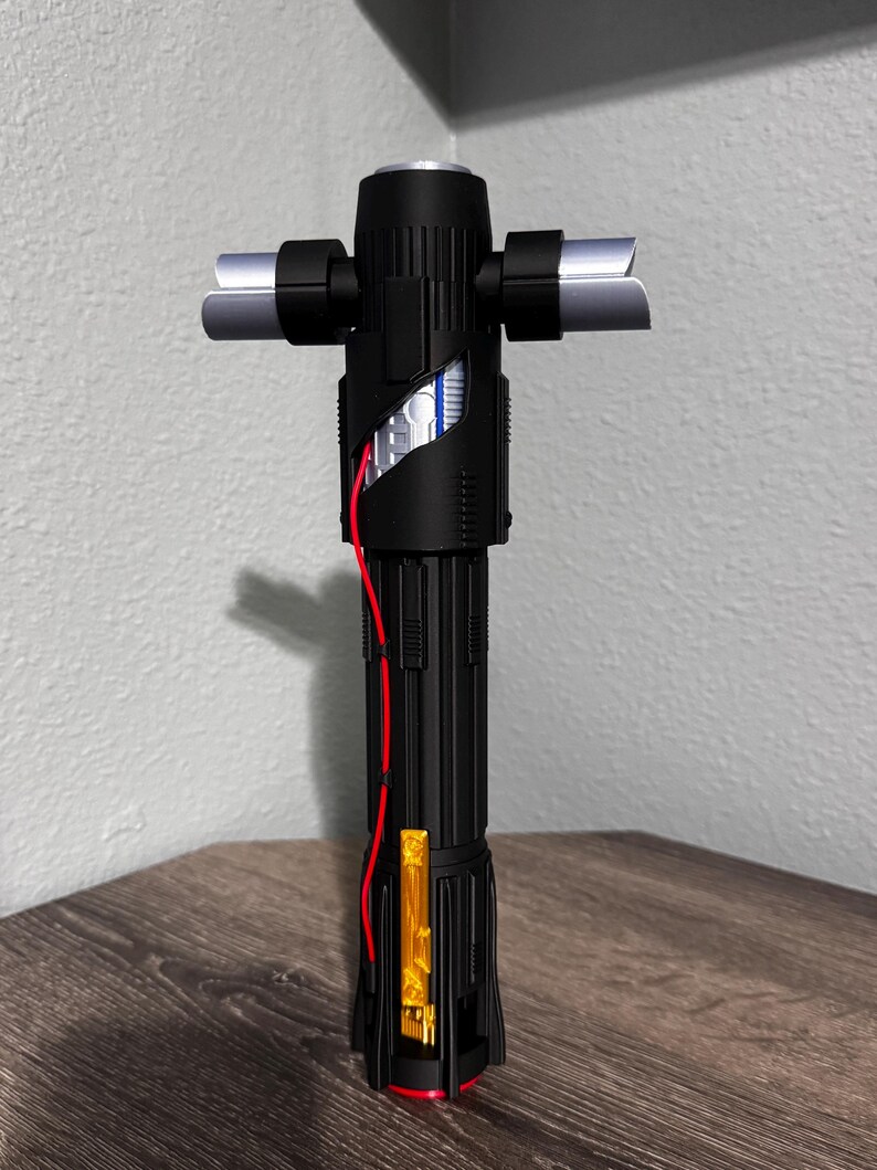 May include: A black lightsaber with silver crossguard blades and a red wire detail. The hilt has a cylindrical design with a gold and red accent. The lightsaber is displayed on a wooden surface.