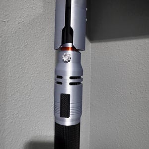 Puede incluir: Una empu&ntilde;adura de sable de luz plateada y negra con un agarre texturizado. La empu&ntilde;adura tiene un dise&ntilde;o cil&iacute;ndrico con un acabado plateado y detalles en negro. La secci&oacute;n superior tiene una cubierta plateada. El sable de luz es un accesorio o coleccionable.