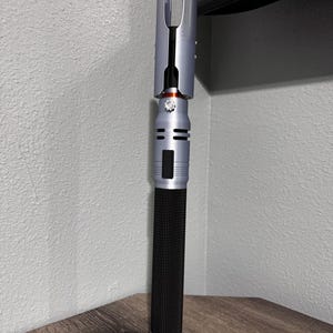Puede incluir: Un sable de luz plateado y negro con un mango negro texturizado. El mango tiene un acabado plateado con un acento rojo y un emisor detallado. El sable de luz se exhibe sobre una superficie de madera.