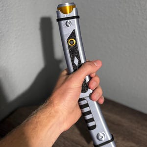 Pu&ograve; includere: Un'elica di spada laser argento e nera con dettagli dorati. L'impugnatura presenta un'impugnatura strutturata, bande nere e un design a forma di diamante. Elastici neri fissano la parte superiore e inferiore.