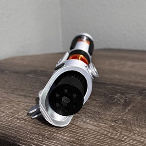 May include: A silver and black lightsaber prop with orange and black detailing. The prop features a detailed design with a cylindrical handle and a black emitter. The lightsaber is angled towards the viewer, highlighting its intricate details.