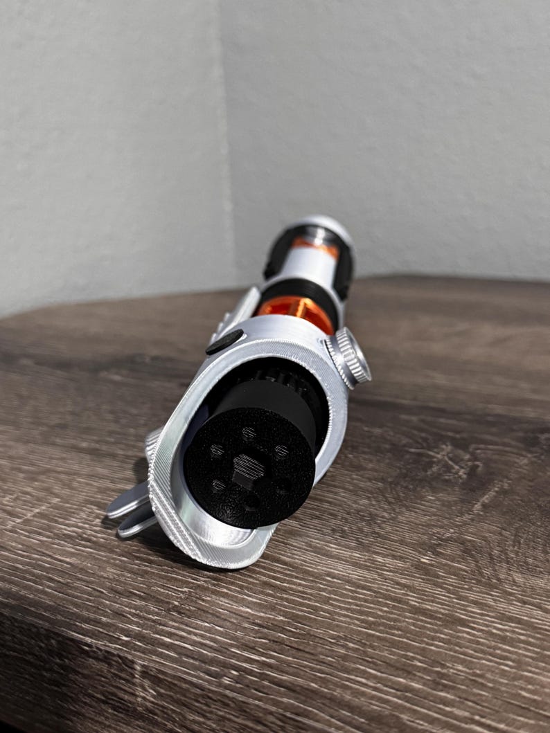 May include: A silver and black lightsaber prop with orange and black detailing. The prop features a detailed design with a cylindrical handle and a black emitter. The lightsaber is angled towards the viewer, highlighting its intricate details.