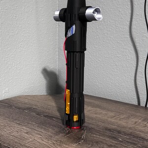 May include: A black lightsaber with silver detailing and a crossguard. The hilt has a ribbed design, with a red accent near the base. The lightsaber is displayed on a wooden surface.