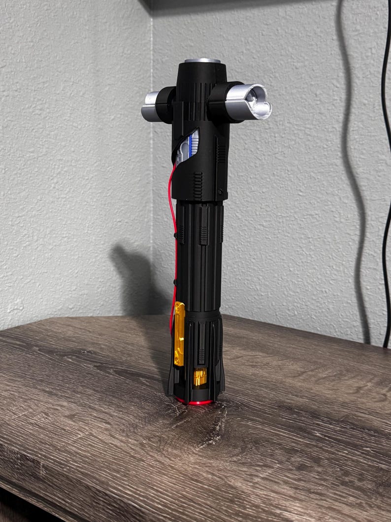 May include: A black lightsaber with silver detailing and a crossguard. The hilt has a ribbed design, with a red accent near the base. The lightsaber is displayed on a wooden surface.