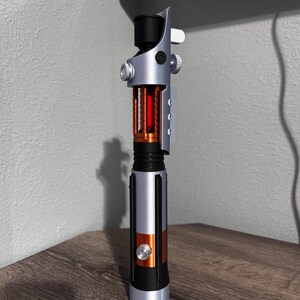 May include: A silver and black lightsaber with orange and red detailing. The hilt has a cylindrical design with a black grip, an orange midsection, and a silver emitter. The blade section has a red interior.
