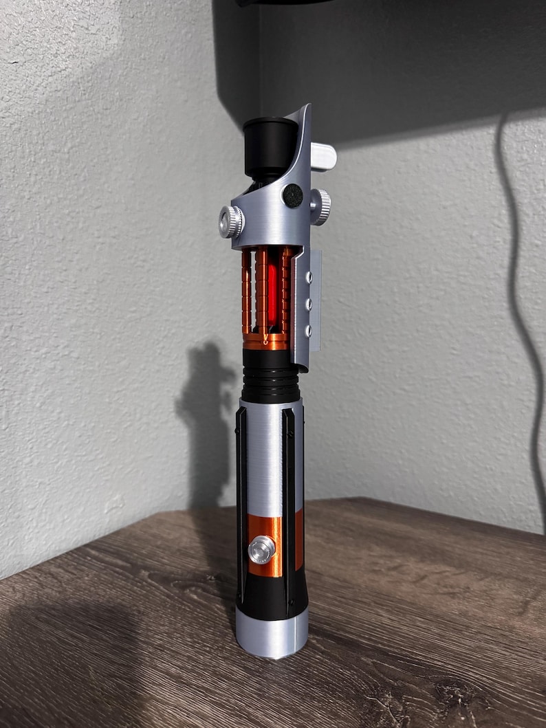 May include: A silver and black lightsaber with orange and red detailing. The hilt has a cylindrical design with a black grip, an orange midsection, and a silver emitter. The blade section has a red interior.