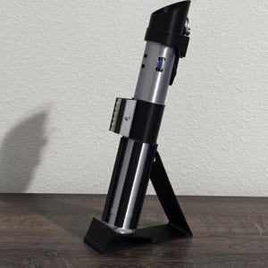 Pu&ograve; includere: Una replica di spada laser argento e nera, esposta su un supporto nero. La spada laser ha un design cilindrico con sezioni dettagliate e un'impugnatura nera. Il supporto &egrave; angolato, fornendo supporto per l'accessorio.