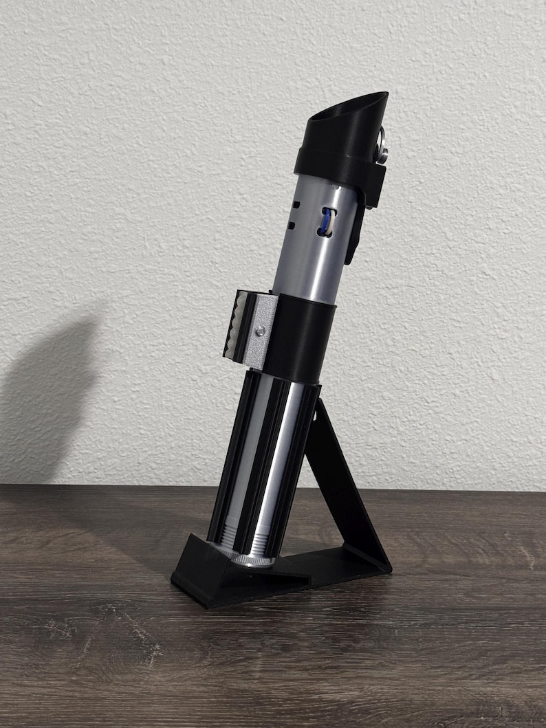 Pu&ograve; includere: Una replica di spada laser argento e nera, esposta su un supporto nero. La spada laser ha un design cilindrico con sezioni dettagliate e un'impugnatura nera. Il supporto &egrave; angolato, fornendo supporto per l'accessorio.