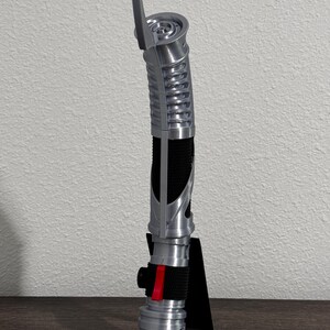May include: A silver and black lightsaber prop, featuring a ribbed handle and a red accent. The prop is displayed on a black stand, showcasing its detailed design. The lightsaber has a curved design and a blade emitter.