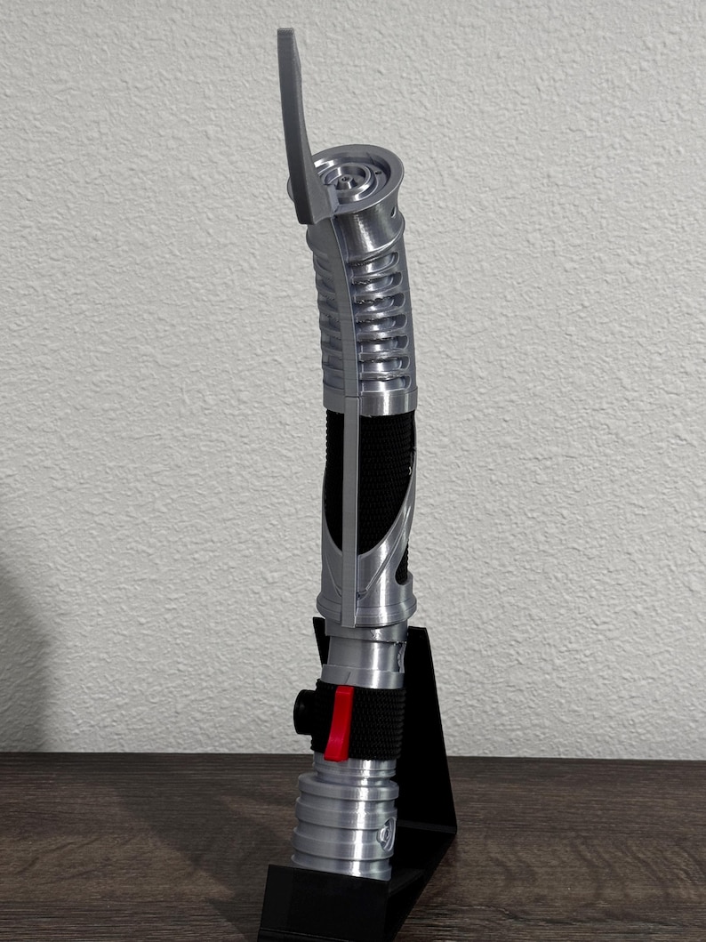 May include: A silver and black lightsaber prop, featuring a ribbed handle and a red accent. The prop is displayed on a black stand, showcasing its detailed design. The lightsaber has a curved design and a blade emitter.