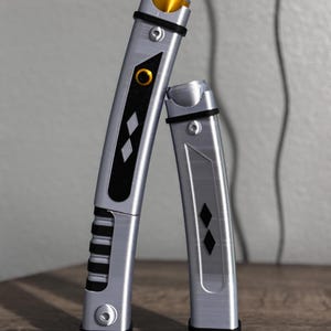Pu&ograve; includere: Due spade laser argentate e nere con dettagli dorati. Le spade laser hanno un design rettangolare con forme a diamante nere e un dettaglio circolare dorato. Le impugnature hanno un design a coste. Le spade laser sono appoggiate a un muro grigio.