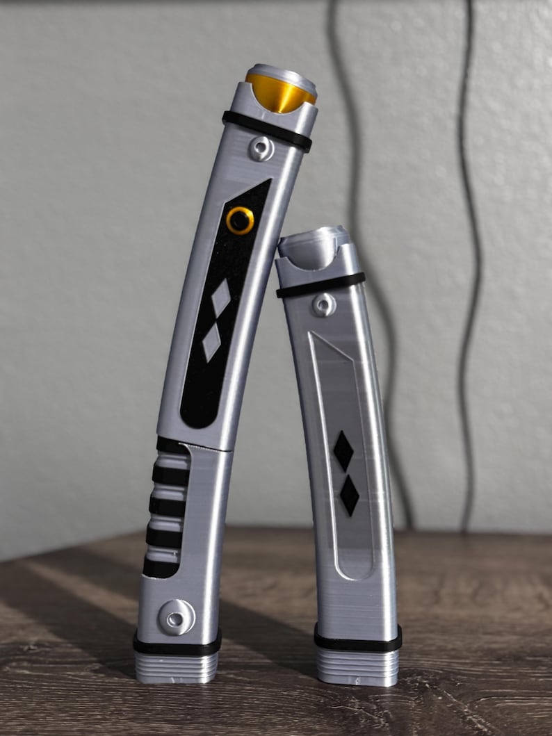 Pu&ograve; includere: Due spade laser argentate e nere con dettagli dorati. Le spade laser hanno un design rettangolare con forme a diamante nere e un dettaglio circolare dorato. Le impugnature hanno un design a coste. Le spade laser sono appoggiate a un muro grigio.