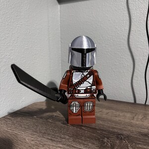 May include: A plastic toy figure featuring a silver helmet and brown armor with white and black detailing. The figure holds a black sword in its right hand and has a blocky, articulated design with a T-shaped visor on the helmet.