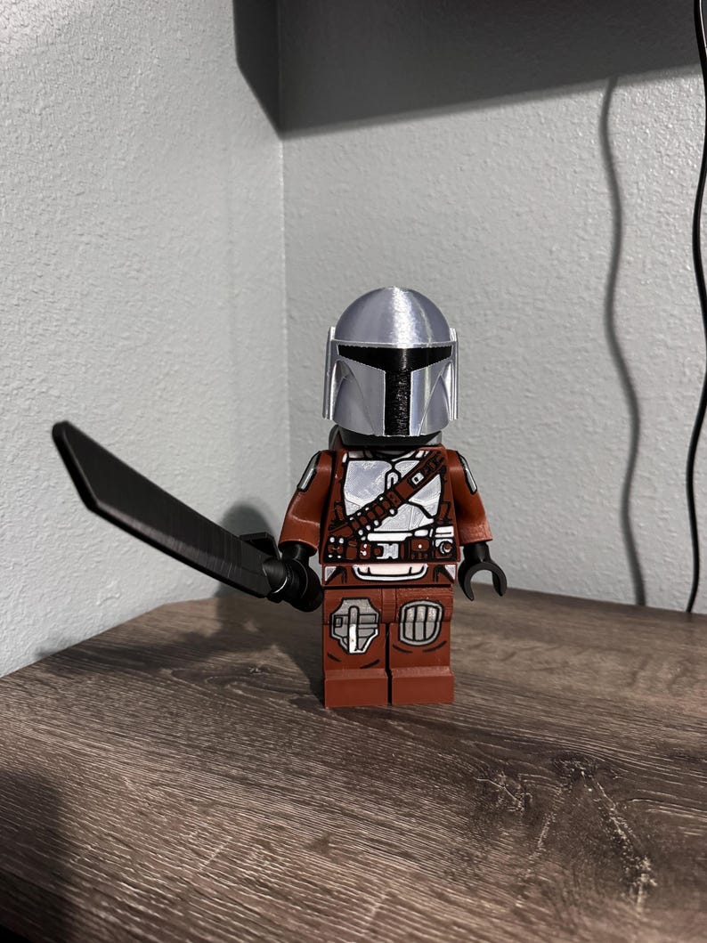 May include: A plastic toy figure featuring a silver helmet and brown armor with white and black detailing. The figure holds a black sword in its right hand and has a blocky, articulated design with a T-shaped visor on the helmet.