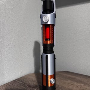 May include: A lightsaber prop in silver, black, and orange. The hilt has a cylindrical design with a red glowing core. A silver button and accents are visible. The lightsaber is displayed on a wooden surface.