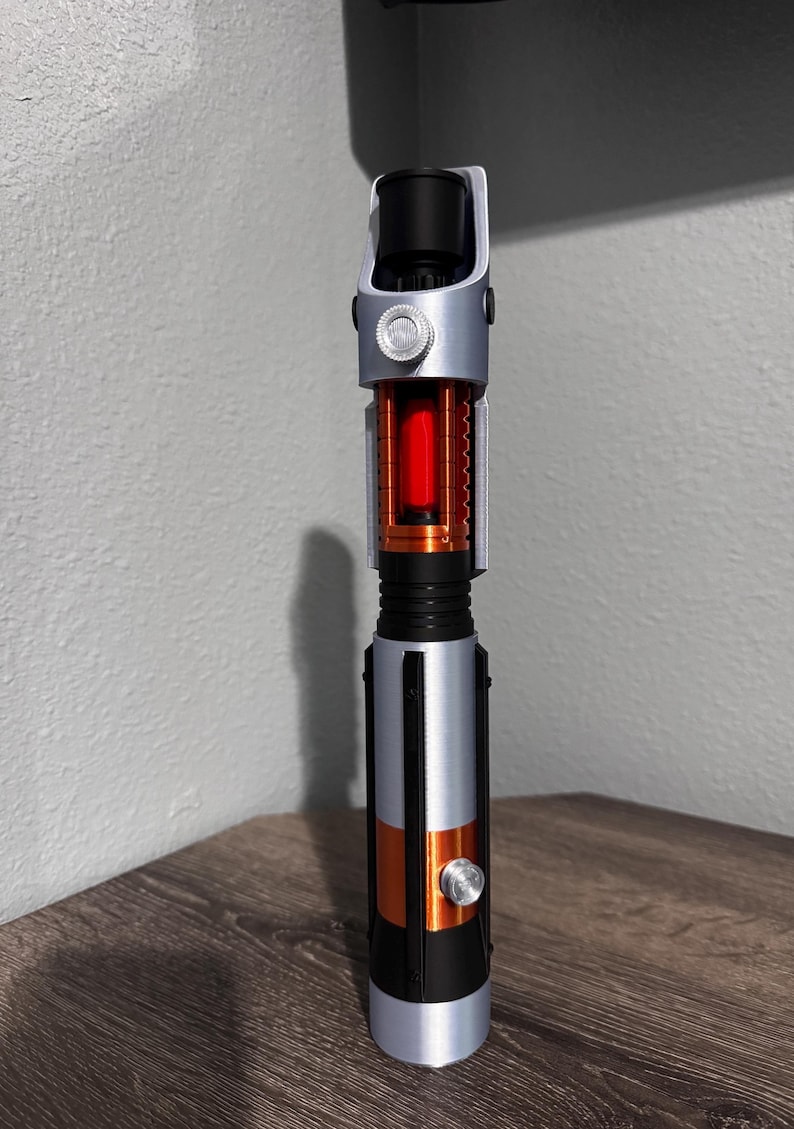 May include: A lightsaber prop in silver, black, and orange. The hilt has a cylindrical design with a red glowing core. A silver button and accents are visible. The lightsaber is displayed on a wooden surface.