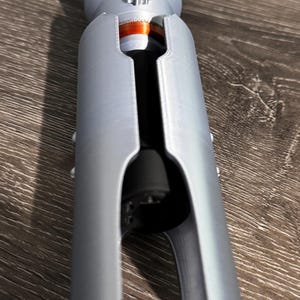 Puede incluir: Una empu&ntilde;adura de sable de luz plateada con un dise&ntilde;o detallado. La empu&ntilde;adura tiene forma cil&iacute;ndrica con un agarre texturizado y una secci&oacute;n de emisor prominente. La abertura de la hoja revela un interior negro y un acento rojo.