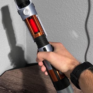 May include: A lightsaber prop with a silver, black, and orange design. The hilt has a cylindrical shape with detailed features, including a visible red energy core. The prop is held against a neutral grey background.