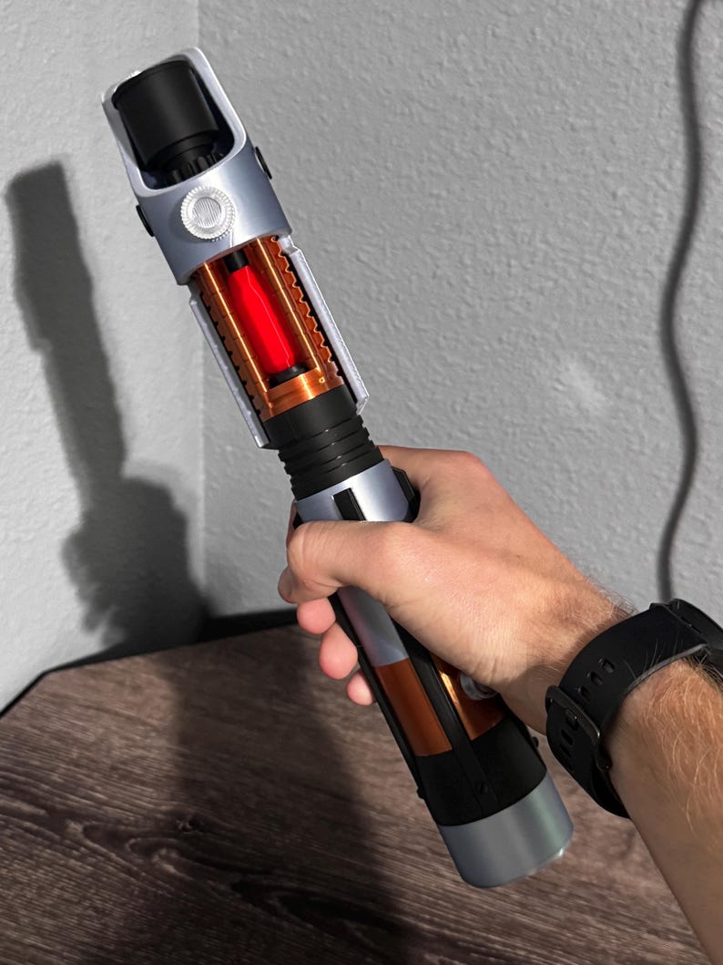 May include: A lightsaber prop with a silver, black, and orange design. The hilt has a cylindrical shape with detailed features, including a visible red energy core. The prop is held against a neutral grey background.