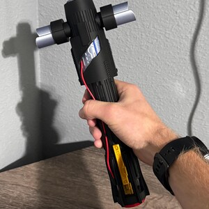 May include: Black lightsaber with silver crossguard blades. The hilt has a detailed design with red and yellow accents. The lightsaber is held in a hand, with a black watch visible on the wrist.