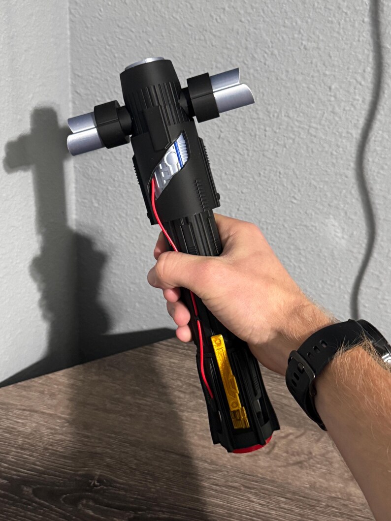 May include: Black lightsaber with silver crossguard blades. The hilt has a detailed design with red and yellow accents. The lightsaber is held in a hand, with a black watch visible on the wrist.