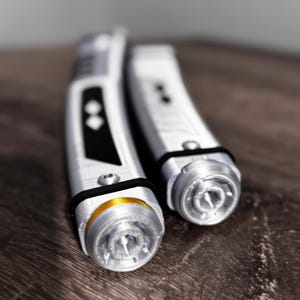 Pu&ograve; includere: Due spade laser argento e nere con dettagli intricati. Una ha un accento dorato vicino all'emettitore. Gli elsi sono cilindrici con fasce nere. Le spade laser sono su una superficie di legno scuro.