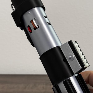 Pu&ograve; includere: Un'elica di spada laser argento e nera con manico e dettagli neri. La spada laser ha un corpo cilindrico con una serie di fori e un emettitore nero. L'elica &egrave; tenuta in mano.