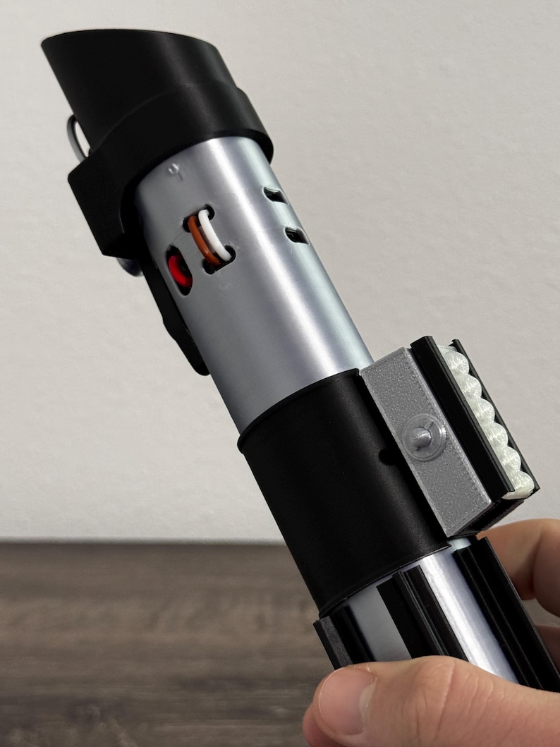 Pu&ograve; includere: Un'elica di spada laser argento e nera con manico e dettagli neri. La spada laser ha un corpo cilindrico con una serie di fori e un emettitore nero. L'elica &egrave; tenuta in mano.