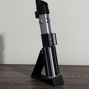 Pu&ograve; includere: Una replica di spada laser nera e argento su un supporto nero. La spada laser ha un emettitore nero, corpo argentato e un'impugnatura nera con dettagli argento e neri. Il supporto &egrave; un design semplice e angolato.