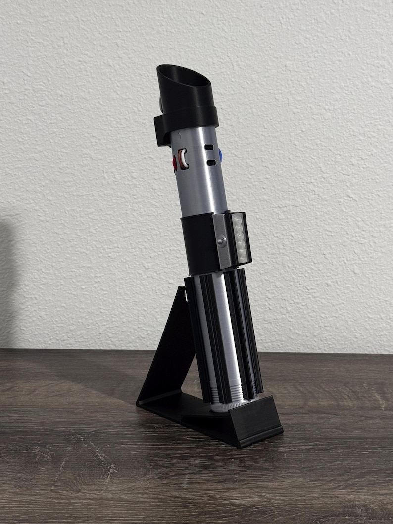 Pu&ograve; includere: Una replica di spada laser nera e argento su un supporto nero. La spada laser ha un emettitore nero, corpo argentato e un'impugnatura nera con dettagli argento e neri. Il supporto &egrave; un design semplice e angolato.